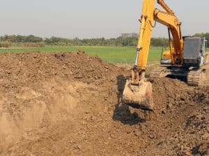 Operate excavator digging large dirt pit in a field with green landscape background.