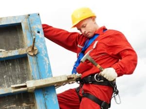 Construction worker in safety gear with a white card certificate, climbing a scaffold while holding a hammer.