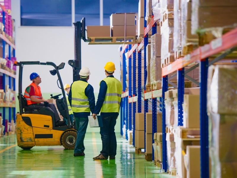 Forklift operator lifting boxes while supervisors observe, showcasing forklift safety in warehouse operations.