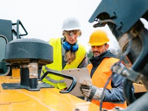 Two workers in safety gear discussing a workplace first aid plan near heavy machinery for risk assessment.