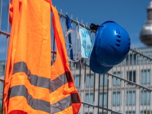 High-visibility vest, safety helmet, and mask on a fence emphasising site risk assessment and proper safety measures.