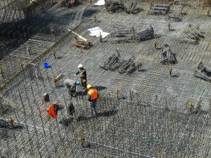 Workers on rebar grid, white card required for construction site access. High angle view, safety vests, and building materials visible.